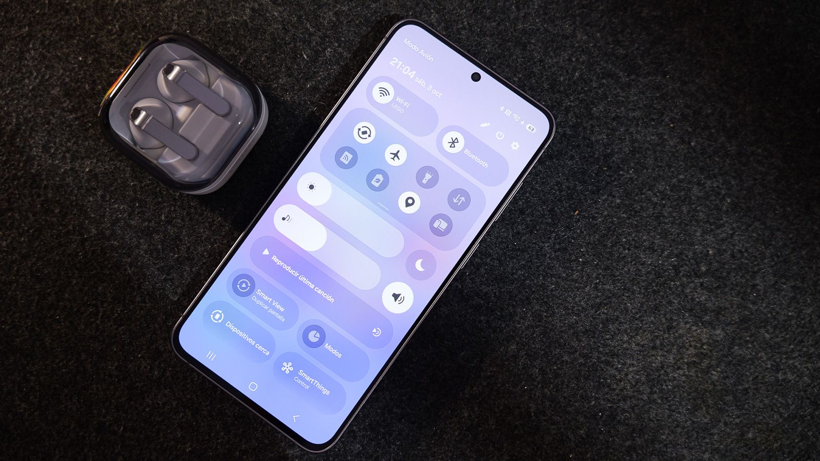Teléfono móvil con el panel de ajustes rápidos de la interfaz One UI desplegado junto a unos auriculares inalámbricos sobre una superficie oscura.
