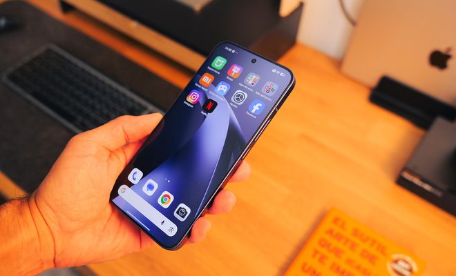 Mano sujetando un Xiaomi 17T Pro encendido sobre una mesa de madera con teclado y portátil al fondo.
