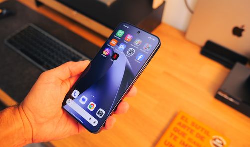 Mano sujetando un Xiaomi 17T Pro encendido sobre una mesa de madera con teclado y portátil al fondo.