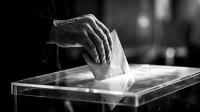 Primer plano en blanco y negro de una mano introduciendo un sobre en una urna electoral transparente.