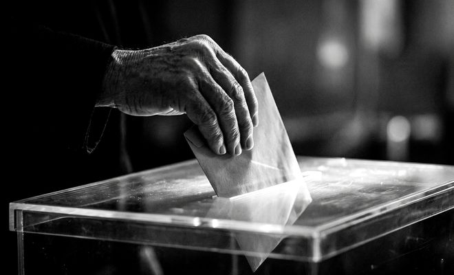 Primer plano en blanco y negro de una mano introduciendo un sobre en una urna electoral transparente.