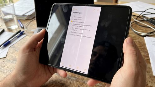 Manos sosteniendo un dispositivo móvil de gran formato abierto que muestra una aplicación de notas sobre un escritorio de trabajo.