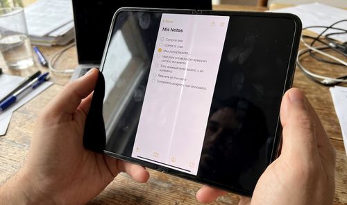 Manos sosteniendo un dispositivo móvil de gran formato abierto que muestra una aplicación de notas sobre un escritorio de trabajo.