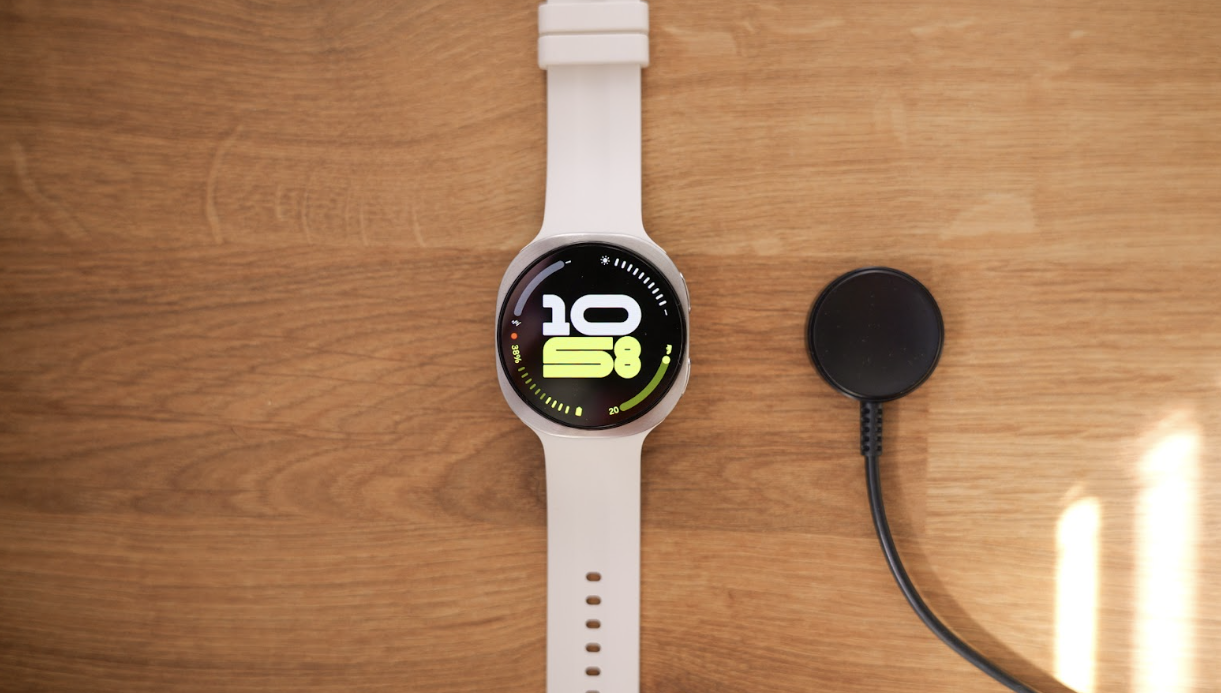Vista cenital de un reloj inteligente Samsung Galaxy Watch9 con correa blanca junto a su base de carga magnética sobre una mesa de madera.