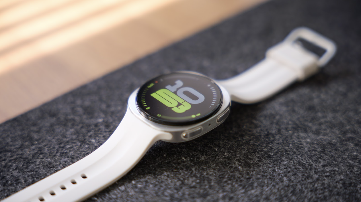 Perspectiva diagonal del Samsung Galaxy Watch9 con correa blanca y esfera digital verde sobre una alfombrilla gris.