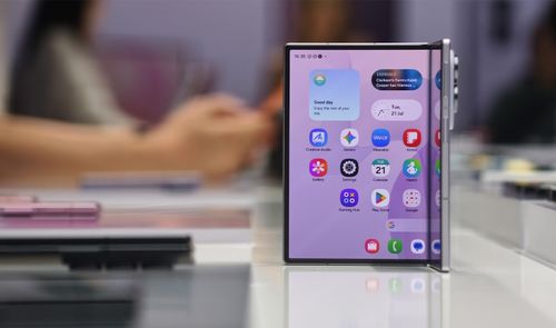 Smartphone plegable Samsung Galaxy Z-Fold8 apoyado verticalmente sobre una mesa blanca mostrando su pantalla principal con iconos de aplicaciones y widgets.