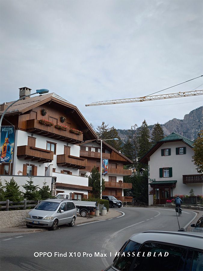 Calle de un pueblo alpino con casas de madera, un coche aparcado y montañas al fondo bajo un cielo nublado.