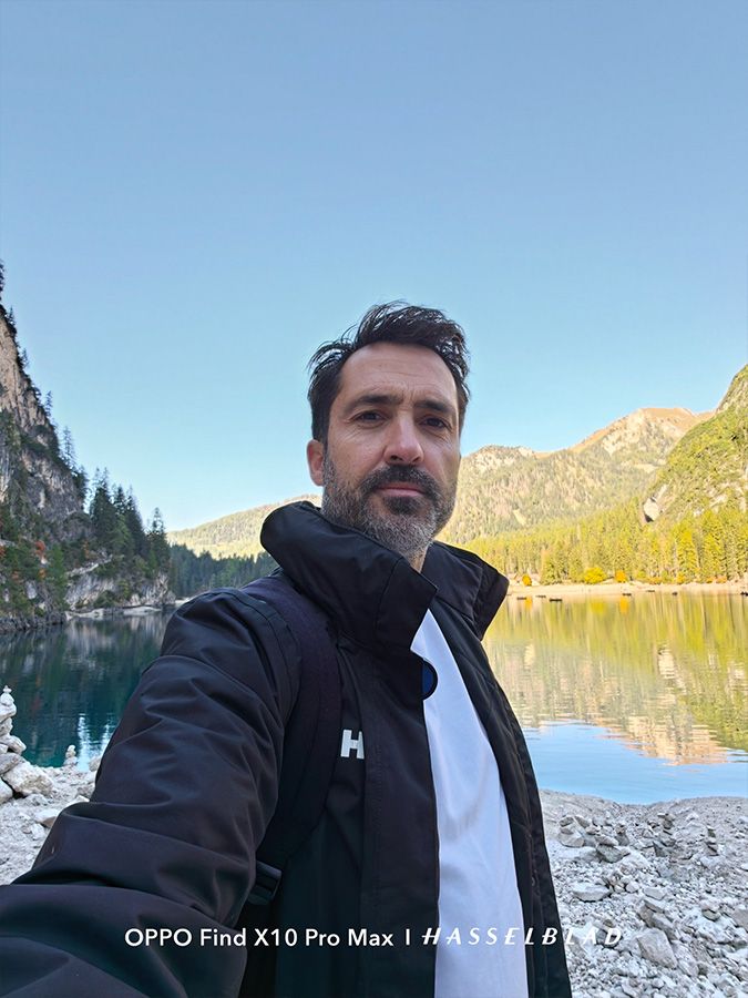 Primer plano de un hombre tomándose un selfie en un entorno natural con montañas, un lago y cielo despejado.