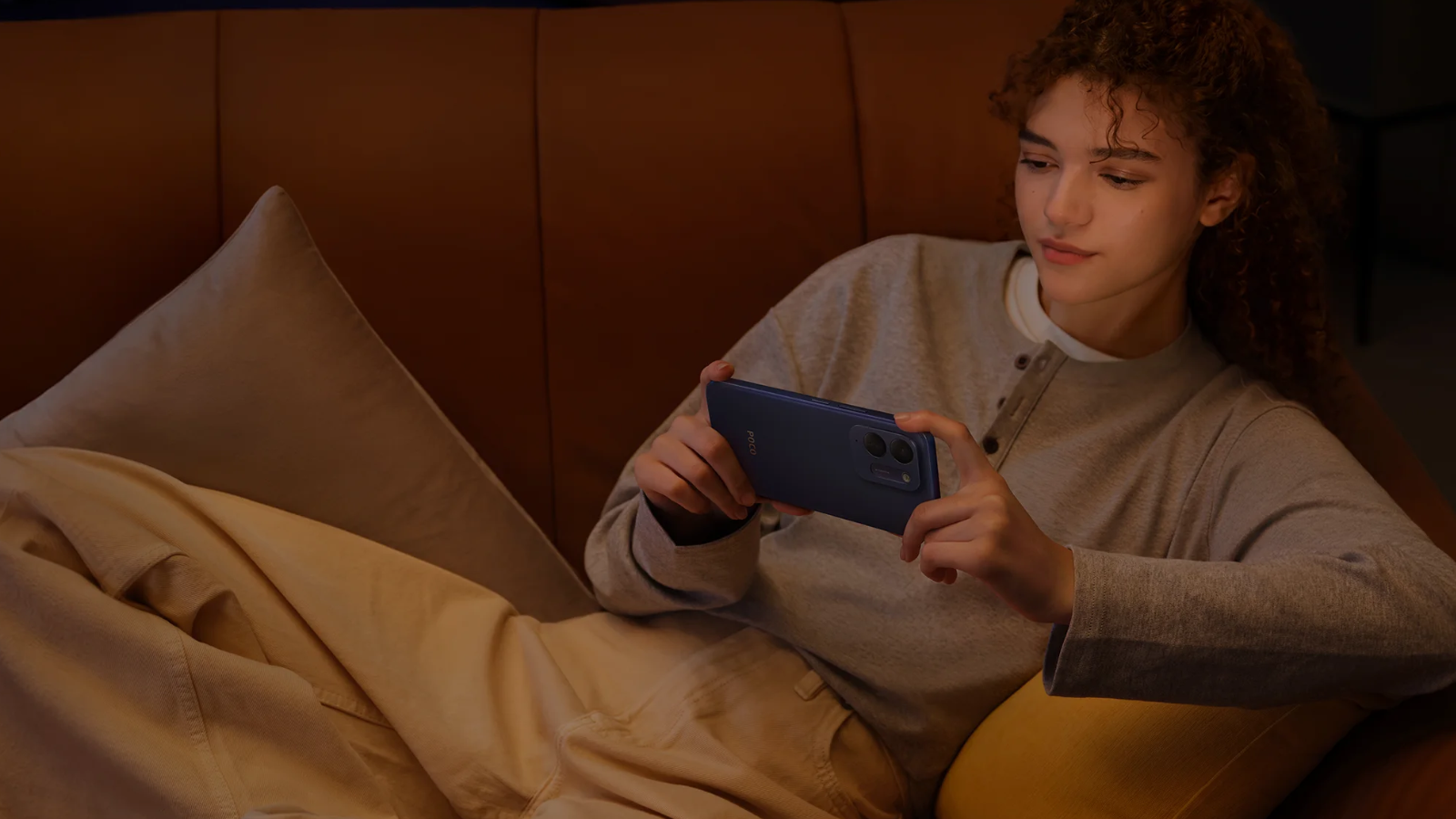 Mujer joven sentada en un sofá sosteniendo horizontalmente el teléfono POCO C95 Pro en color azul.