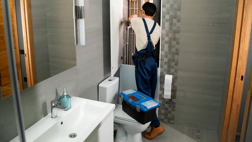 Fontanero de espaldas manipulando una instalación tras un panel en un cuarto de baño con una caja de herramientas azul sobre el inodoro.