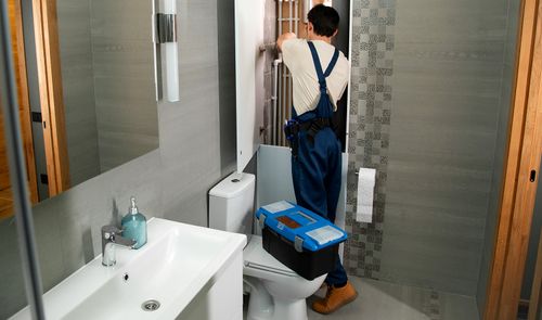 Fontanero de espaldas manipulando una instalación tras un panel en un cuarto de baño con una caja de herramientas azul sobre el inodoro.
