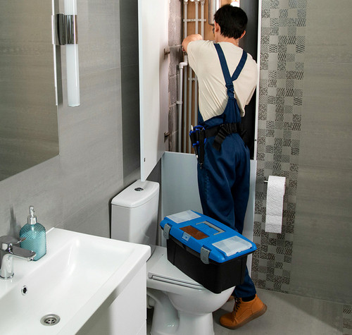 Fontanero de espaldas manipulando una instalación tras un panel en un cuarto de baño con una caja de herramientas azul sobre el inodoro.