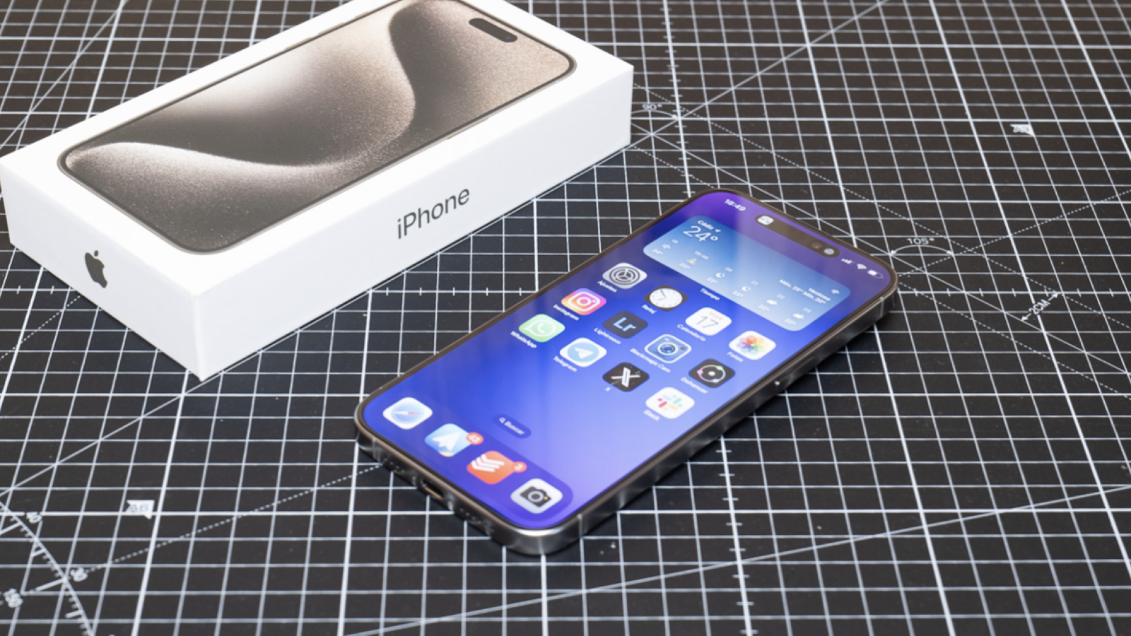 iPhone 15 Pro con la pantalla encendida junto a su caja sobre una base de corte cuadriculada.