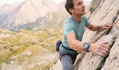 Hombre escalando una pared de roca en la montaña con un reloj inteligente deportivo en su muñeca.