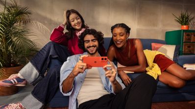 Tres personas sentadas en un sofá mirando la pantalla de un teléfono moto g06 de color rojo en posición horizontal.