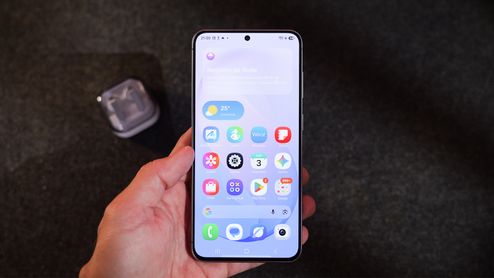 Mano sujetando un teléfono móvil Samsung con la pantalla de inicio de One UI 9 donde se aprecian iconos de aplicaciones y un widget meteorológico.