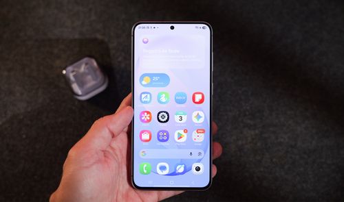 Mano sujetando un teléfono móvil Samsung con la pantalla de inicio de One UI 9 donde se aprecian iconos de aplicaciones y un widget meteorológico.