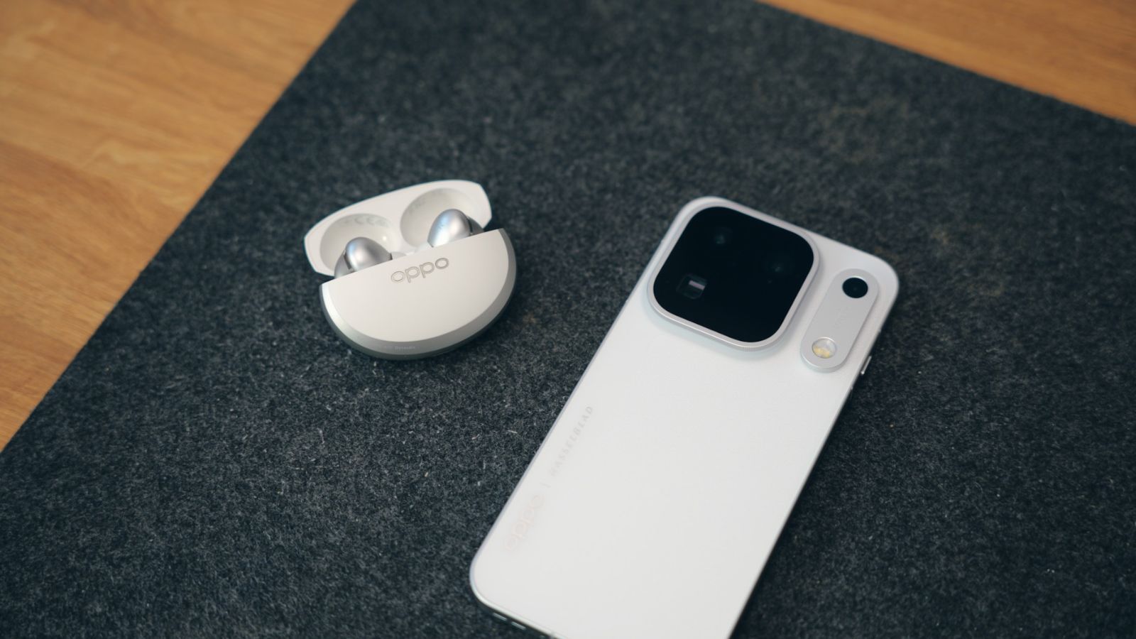 Auriculares OPPO Enco X4 en su estuche de carga junto a la parte trasera de un smartphone blanco sobre una superficie gris.