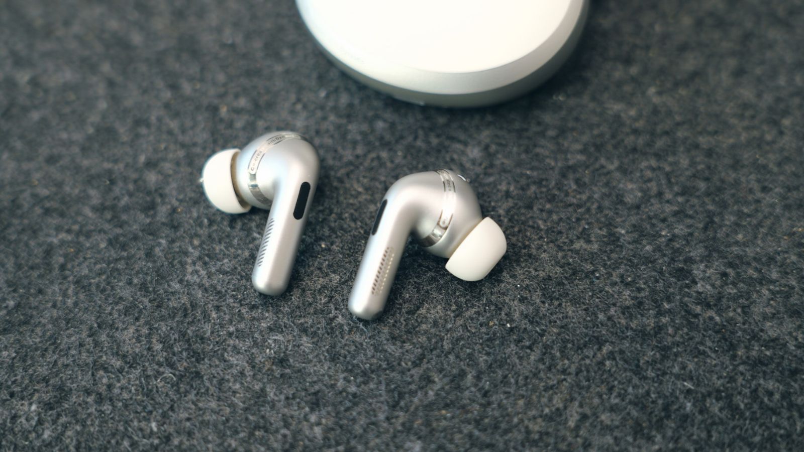 Primer plano de los auriculares OPPO Enco X4 de color plateado con almohadillas blancas sobre una base textil oscura.