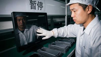 Operario con uniforme y guantes blancos inspeccionando la pantalla de un monitor frente a una línea de montaje de teléfonos móviles.
