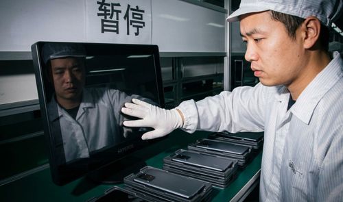 Operario con uniforme y guantes blancos inspeccionando la pantalla de un monitor frente a una línea de montaje de teléfonos móviles.