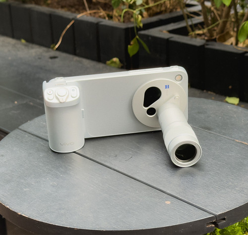 Teléfono vivo-X500 Pro Max con accesorio de teleconvertidor óptico y empuñadura fotográfica sobre una mesa exterior.