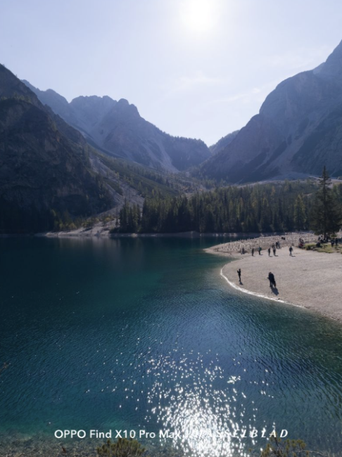 Lago de montaña con aguas turquesas y reflejos del sol junto a una orilla de arena con personas.