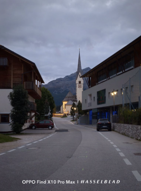 Calle de un pueblo alpino al anochecer con una iglesia de torre afilada al fondo bajo un cielo nublado.