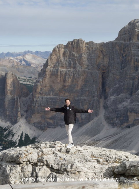 Hombre posando con los brazos abiertos en la cima de una montaña rocosa frente a grandes macizos de piedra.