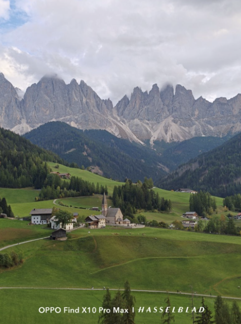 Pequeño pueblo de montaña con iglesia rodeado de prados verdes bajo una cordillera de picos escarpados.